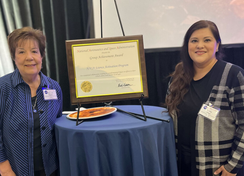 Image of Margaret Baguio and Celena Miller standing next to NASA Group Achievement Award that is displayed on a table.